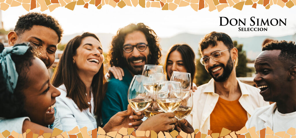 A diverse group of friends smiles and clinks glasses of white wine outdoors at sunset, celebrating together. The words Don Simon Seleccion appear in the top right corner with a mosaic border.